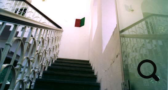 Treppe mit weißen Geländern, die nach oben zu einem hellen Raum führt; an der Wand hängt eine rot-grüne Flagge.